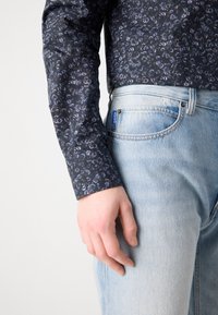 Camicia a maniche lunghe in tessuto blu scuro con un motivo floreale, abbinata a jeans in denim azzurro chiaro con un'etichetta visibile sulla tasca.