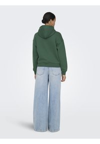 Femme avec les cheveux en chignon, portant un sweat à capuche vert et un jean large bleu clair, debout de dos devant un fond clair uni.