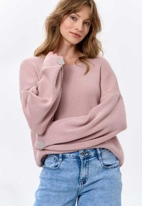 Hellrosa gerippter Pullover mit weitem Ausschnitt, übergroßen Ärmeln und Knopfdetails an den Bündchen, kombiniert mit hellblauen Jeans.