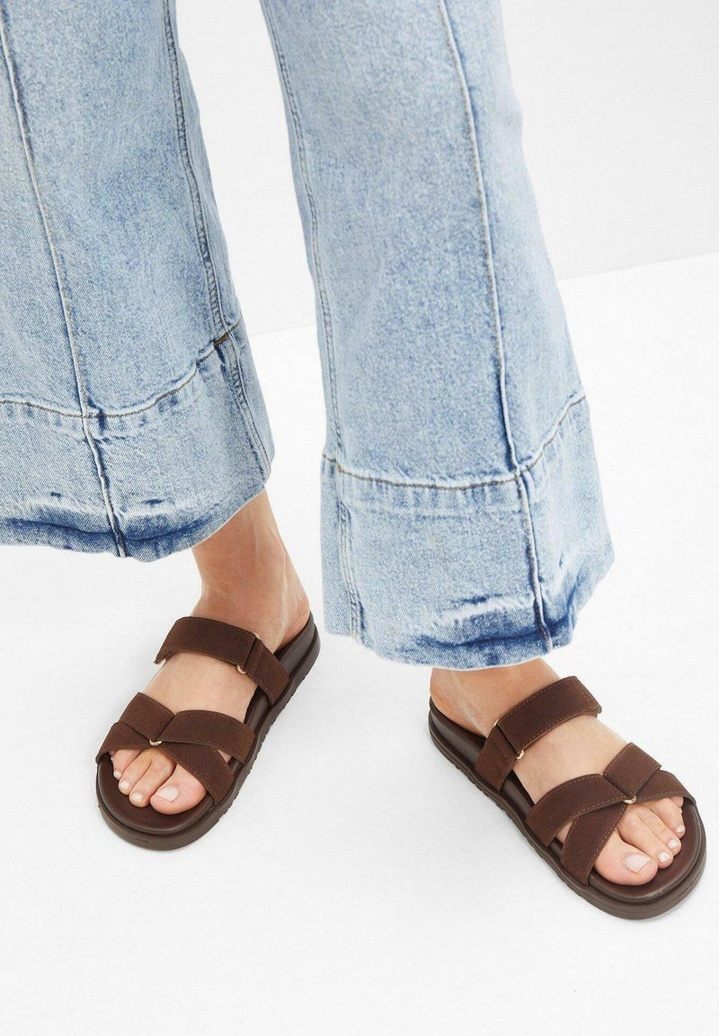 Sandales marron avec deux larges brides et un design croisé. Semelle épaisse avec un détail texturé. Associées à un jean évasé bleu clair à longueur cheville.