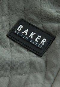 Kviltet grå stof med en glat tekstur, der har et sort rektangulært mærke med præget hvide bogstaver, der står "BAKER BY TED BAKER."