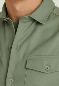 Lichtgroen shirt met een kraag, voorzien van twee voorzakken en knoopsluitingen. Gladde stofstructuur met een matte afwerking.