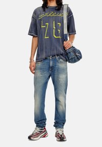 Camisa de mezclilla azul de corte oversize con letras amarillas, combinada con jeans claros desgastados y zapatillas con acentos rojos. Lleva una bolsa de mezclilla azul.