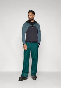 Χιονοδρομικό μπουφάν σε σκούρο γκρι και τιρκουάζ με φερμουάρ εμπρός, αντιθετικές πινελιές και κουκούλα· συνδυασμένο με τιρκουάζ αδιάβροχες παντελόνες και μαύρα παπούτσια σκι.