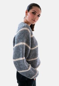 Grijze gebreide trui met witte horizontale strepen, gestructureerde stof, relaxed fit en brede mouwen. G gedragen met zwarte broek.