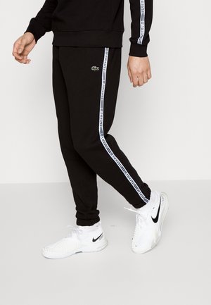 Un pantalon de survêtement noir avec des bandes blanches latérales sur lesquelles un texte est répété, accompagné d'un petit logo Lacoste vert. Associé à des chaussures de sport blanches.