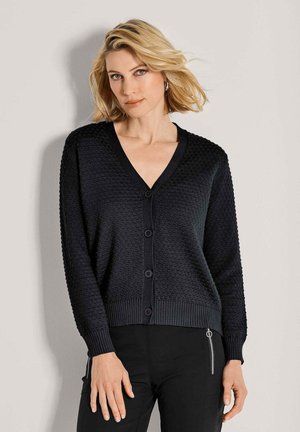 Blonde Frau, die einen strukturierten schwarzen, knopfgebundenen Cardigan und schwarze Hosen mit einem Reißverschluss trägt, steht vor einer hellgrauen Wand.