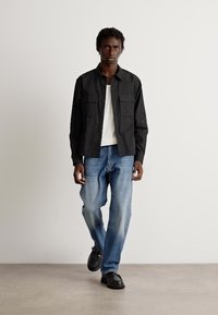 Svart skjorte med knapper og to brystlommer, hvit T-skjorte under, falmede blå jeans og svarte sko. Modell går på en nøytral bakgrunn.