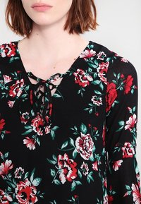 Musta kukkatuote, jossa on punaisia ja valkoisia kukkia, V-pääntie nauhakiinnityksellä ja pitkät hihat. Kevyt, teksturoitu kangas, joka on rento istuvuus.
