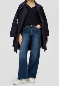 Marinblå trenchcoat över en svart v-ringad tröja, kombinerat med vida blå jeans och vita sneakers med plattform. Noterbara detaljer med knappar och fickor.