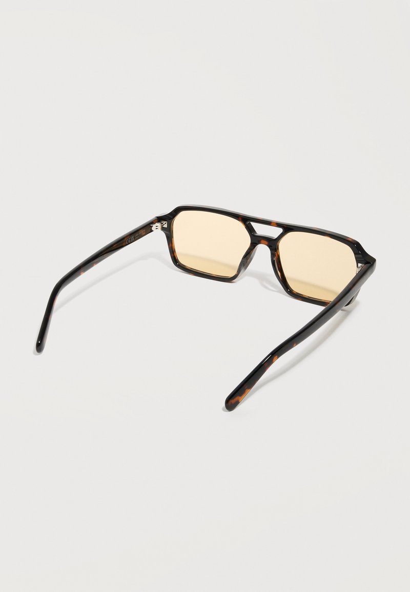 Lunettes de soleil rectangulaires en écaille de tortue avec des verres teintés ambre, posées à plat sur une surface blanche.