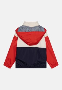 Fargeblokkert vindjakke med røde, marineblå og kremfargede paneler, stripete ryggstykke, elastiske mansjetter og hette med snøring.