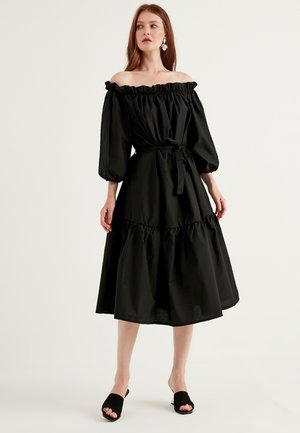 Abito nero off-the-shoulder con scollatura voluminosa, maniche lunghe a palloncino, vita cinchata con fiocco e gonna a strati svasata. Tessuto di cotone liscio.