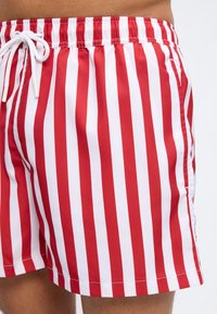 Κόκκινα και λευκά κατακόρυφα ριγέ μαγιό βερμούδα με λευκό κορδόνι στη μέση και το πάνω μέρος του μηρού ενός ατόμου, πάνω σε απλό φόντο.