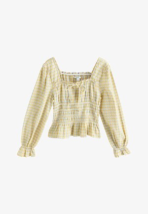 Dzeltens krekls ar garām pūkainām piedurknēm, laukveida kakla izgriezumu un auklas detaļu. Raksturots ar savāktu tekstūru un volānu apakšmalu.