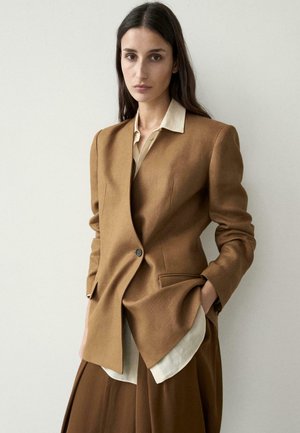 Donna con lunghi capelli scuri indossa un blazer marrone su misura sopra una camicia beige, in piedi contro uno sfondo chiaro e uniforme con una mano in tasca.