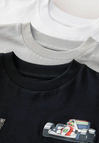 Tre t-shirt a manica corta piegate, di colore bianco, grigio chiaro e nero; la maglietta nera presenta una grafica colorata di una macchina da corsa vicino al collo.