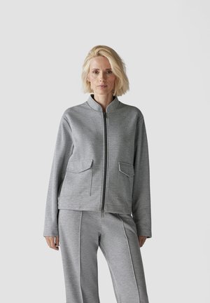 Graue Zip-Jacke mit zwei Fronttaschen, strukturiertem Stoff, hohem Kragen und passenden grauen Hosen, die einen abgestimmten Look schaffen.