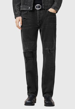 Black denim jeans with distressed details, featuring patchwork, visible stitching, and a relaxed fit. Worn with black boots.