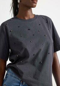 Kortærmet, mørkegrå t-shirt med metalliske dekorationer i sort og sølv, med en afslappet pasform og glat tekstur.