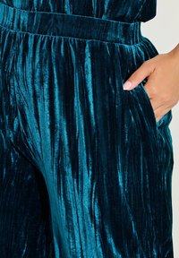 Teal fluwelen broek met verticale ribbeltextuur, voorzien van een brede tailleband en zijzakken. Glad materiaal met een subtiele glans.