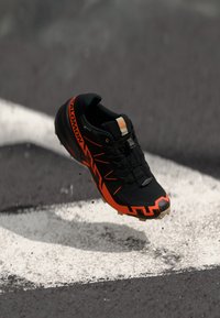 Chaussure de sport noire et orange avec une tige texturée, un système de laçage proéminent et une semelle robuste, présentée en l'air au-dessus d'une ligne blanche.