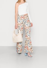 Vit topp med en axel och texturerad tyg, som är kombinerad med blommönstrade, utsvängda byxor i pastellfärger. Håller en ljusbeige handväska.