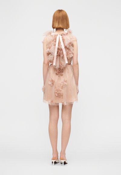 Γυναίκα με κοντά κόκκινα μαλλιά, που φοράει αμάνικο μπεζ φόρεμα από τούλι με λουλουδάτες εφαρμογές και μεγάλο φιόγκο στο πίσω μέρος, συνδυασμένο με λευκά παπούτσια με τακούνι.