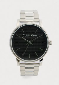 Ρολόι Calvin Klein με μαύρο υφασμάτινο καντράν, διακοσμημένη θήκη και βραχιόλι από ανοξείδωτο ατσάλι σε ασημί απόχρωση και διακριτικούς μεταλλικούς δείκτες.
