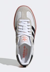 Sneakers Adidas z białej skóry, z szarymi zamszowymi akcentami, czarnymi paskami i gumową podeszwą; zawierają białe sznurowadła i detale logo.