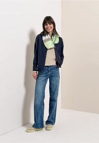 Cardigan marine porté sur un pull beige, jean large bleu, et une écharpe multicolore avec des accents verts et gris. Une chaussure décontractée complète le look.