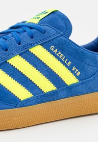 Niebieski zamszowy sneaker z trzema jaskrawymi żółtymi paskami i napisem "GAZELLE VTR" w kolorze złotym. Podeszwa z gumy o teksturowanym wykończeniu.