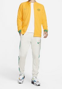 Nike træningsdragt i lys gul med grønne detaljer, indeholdende en jakke med lynlås og en hvid t-shirt, parret med cremede bukser og mønstrede sneakers.