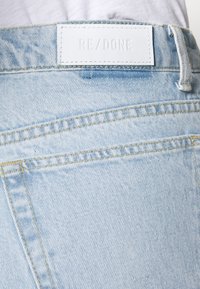 Poche arrière d'un jean en denim bleu clair avec une étiquette rectangulaire blanche cousue au-dessus, affichant le nom de la marque RE/DONE en lettres embossées.