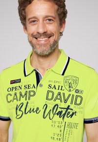 Felgele polo met marineblauwe kraag, voorzien van verschillende geprinte teksten en een logo patch. Glad materiaal en korte mouwen.