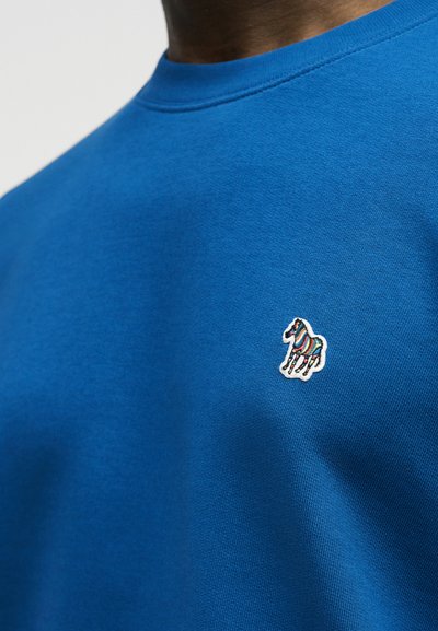 Μπλε πουκάμισο με ένα μικρό κεντητό λογότυπο ενός πολύχρωμου ριγέ ζέβρα στο αριστερό στήθος.