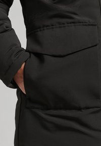Black padded coat featuring a large front pocket, smooth texture, and a snug fit around the wrist, with concealed hand openings.