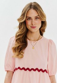 Jasnoróżowa sukienka z bufkami i czerwoną falbaniastą wypustką w pasie. Modelka ma falowane włosy i nałożone warstwowe naszyjniki.