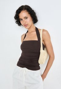 Brown fitted halter top with square neckline, paired with textured beige woven bag. Model wears white loose pants. Smooth fabric, casual style.