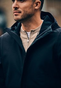 Zwarte capuchonjack gemaakt van gladde stof, met een ritskraag en een beige binnenlaag. Het ontwerp is gestroomlijnd en modern.