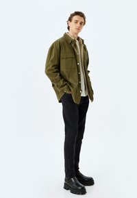 Camisa chaqueta de botón en verde olivo con bolsillos en el pecho, sobre una blusa beige clara, combinada con jeans negros ajustados y botas negras de suela gruesa.