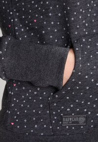 Maglione grigio scuro con piccoli pois bianchi e cuori rosa, con la mano infilata nella tasca anteriore e l'etichetta del marchio Ragwear visibile.