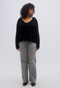 Haut noir à manches longues avec des détails à volants, associé à un jean gris à jambes larges. Le mannequin porte des ballerines noires et a les cheveux bouclés.