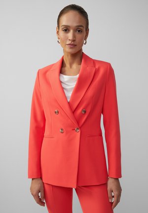Frau trägt einen korallenfarbenen, taillierten Blazer mit doppelter Knopfleiste und passende Hose zu einem weißen Top, steht vor einem einfarbigen hellgrauen Hintergrund.