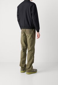 Chaqueta bomber negra con puños acanalados, combinada con pantalones verdes oliva que tienen bolsillos traseros. El calzado es gris con suelas verdes.
