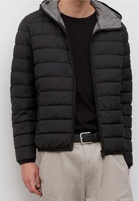 Schwarze Puffjacke mit Reißverschluss, ausgestattet mit einem grauen Kapuzenfutter und gestepptem Design. Getragen über einem schwarzen Shirt mit beigen Hosen.