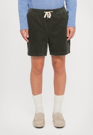 Personne portant un short en velours côtelé vert foncé avec un cordon, des mocassins beige, des chaussettes blanches et une chemise bleue à manches longues.