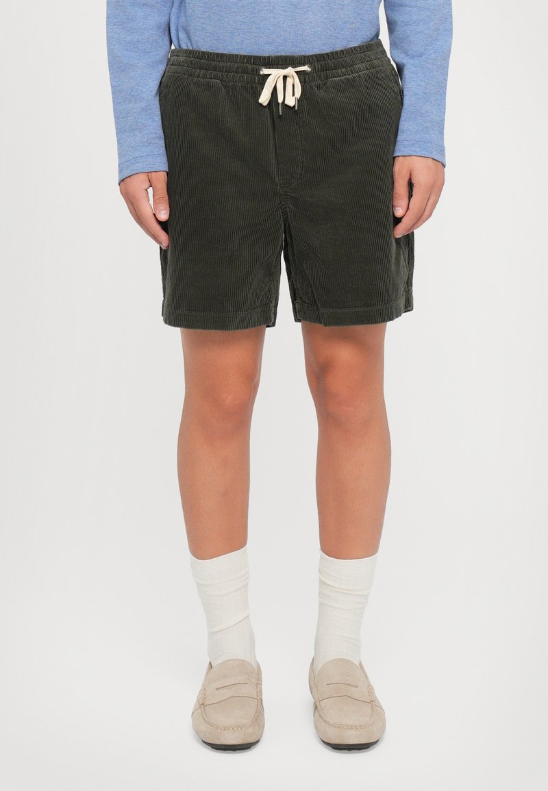 Personne portant un short en velours côtelé vert foncé avec un cordon, des mocassins beige, des chaussettes blanches et une chemise bleue à manches longues.