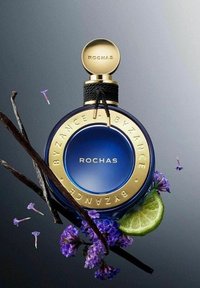Blå rund parfume flaske med guld accenter, der har "ROCHAS" label, omgivet af vaniljestænger, lilla blomster og en lime skive.