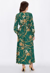 Vestido largo verde con estampado de flores doradas y cadenas, mangas largas, cintura entallada y dobladillo hasta los tobillos. Sandalias doradas de tacón visibles.